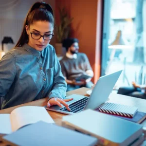 Girl with glasses studying on her laptop at a coffee shop checking her notebook.