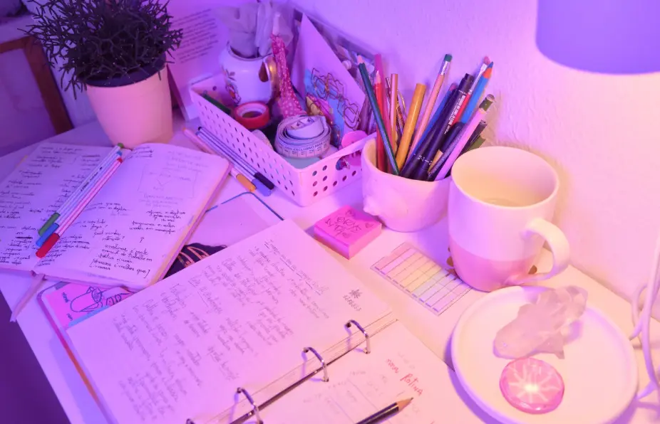 A cozy, well-lit desk setup with notebooks filled with handwritten notes, colorful pens, sticky notes, a cup, a potted plant, and a container holding pencils and markers, all illuminated by a soft purple-pink light.